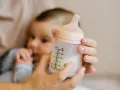 Manos sostienen un biberón con leche, con un bebé borroso al fondo. Es hora de considerar cuando cambiar tetina biberon.