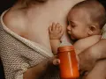 Bebé succionando un biberón naranja, con la mano apoyada en el pecho de la madre. Se aprecian varios tipos de tetinas en el mercado.