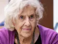 Retrato de una anciana con cabello blanco y ojos azules, luciendo un suéter morado. Su rostro arrugado evoca la sabiduría y el significado de una vida plena, como el nombre Manuela.