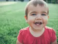 Un bebé de 1 año sin dientes ríe a carcajadas en el césped, mostrando dos pequeños dientes inferiores.