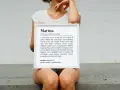Una mujer sostiene un marco blanco con la definición del nombre Marina, que significa "del mar" y "puerto de paz".