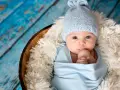 Bebé Ezra, con gorrito azul y envuelto en mantita, mira curioso. Descansa en una cesta sobre fondo de madera azul.
