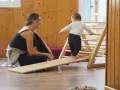 Niña gateando en rampa de madera, animada por mujer sonriente. Fomentando el movimiento libre y la exploración.
