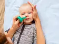 Bebé con ojos azules siendo ayudado a quitar mocos con un aspirador nasal verde.