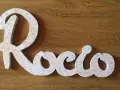Letra decorativa con el nombre Rocio, cubierta de un delicado estampado floral rosa sobre fondo blanco, sobre una superficie de madera.