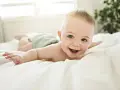 Bebé sonriente boca abajo en una cama blanca, con un reflejo de hociqueo en su expresión juguetona.