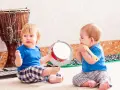 Dos bebés disfrutan de la estimulación musical temprana, uno con un tambor y otro con una flauta, rodeados de instrumentos.