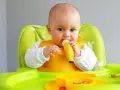 Bebé con babero amarillo comiendo una fruta (plátano) en su trona verde.