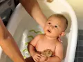 Bebé en bañera, manos adultas ajustan la temperatura del agua. Sostiene una esponja, mirando hacia arriba con curiosidad.
