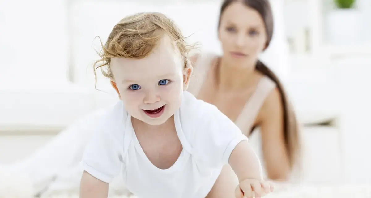 Bebé sonriente gateando, con su madre observando. Una escena tierna para estimular el gateo.
