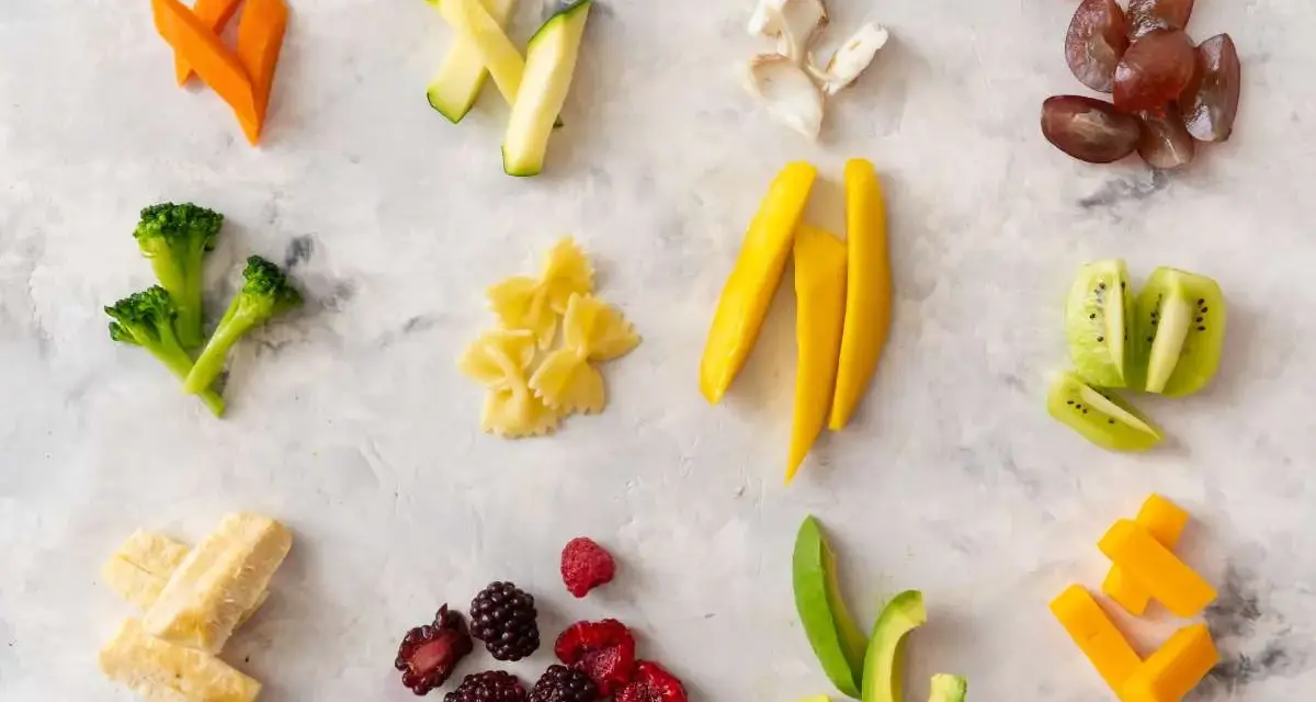 Variedad de alimentos para blw bebé 6 meses: zanahoria, calabacín, brócoli, pasta, mango, kiwi, aguacate, bayas, plátano y queso.