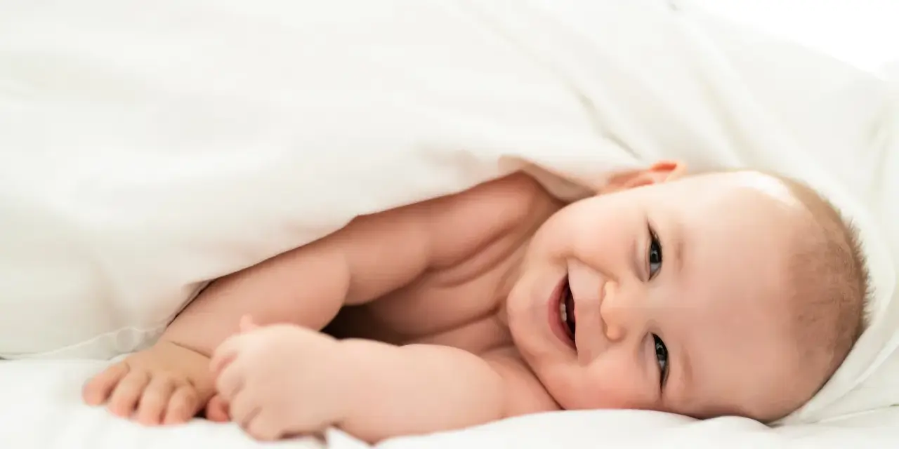 Un bebé sonriente se asoma bajo una manta blanca. Parece que los bebes tienen memoria, ¡y este ya recuerda cómo sonreír!