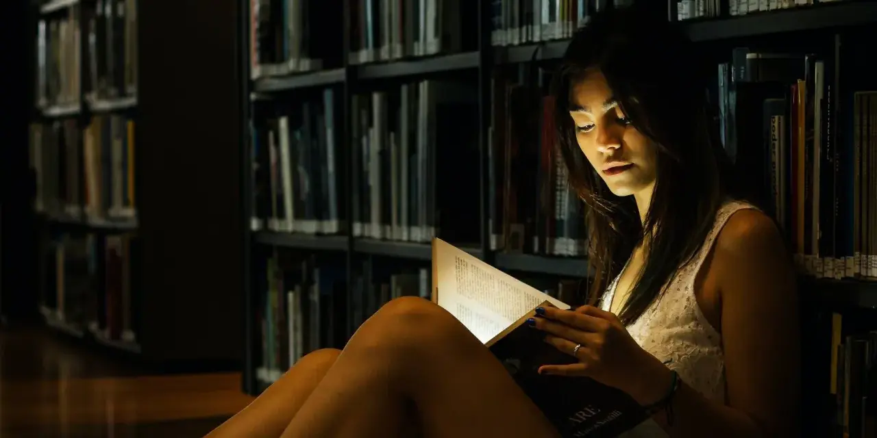 Elena, in the library's dim light, engrossed in a book.