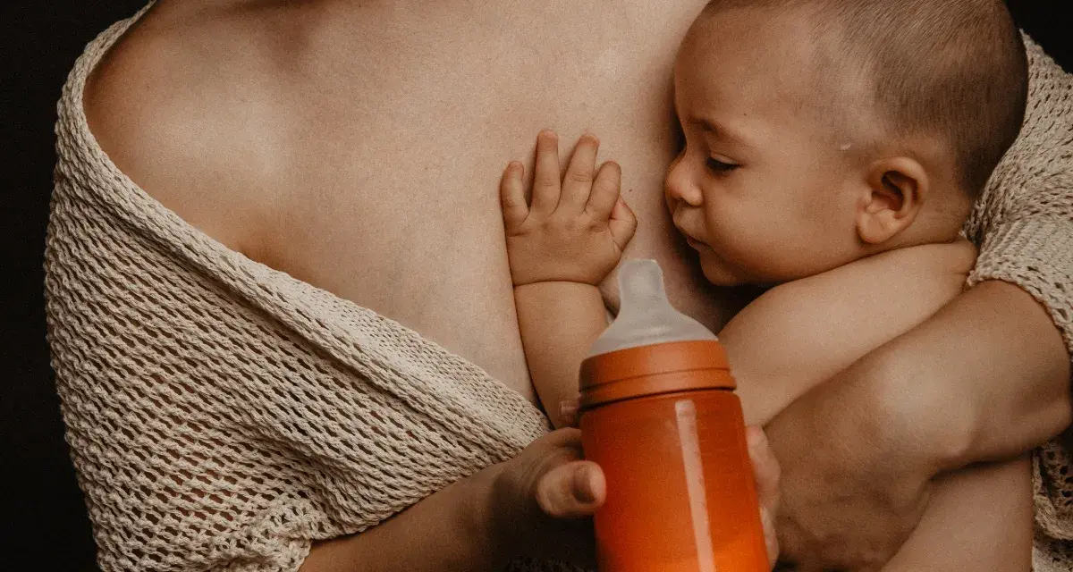 Bebé succionando un biberón naranja, con la mano apoyada en el pecho de la madre. Se aprecian varios tipos de tetinas en el mercado.