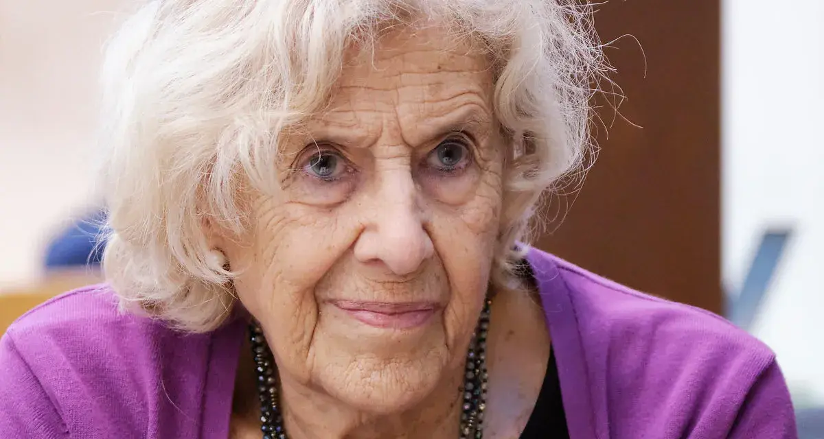 Retrato de una anciana con cabello blanco y ojos azules, luciendo un suéter morado. Su rostro arrugado evoca la sabiduría y el significado de una vida plena, como el nombre Manuela.
