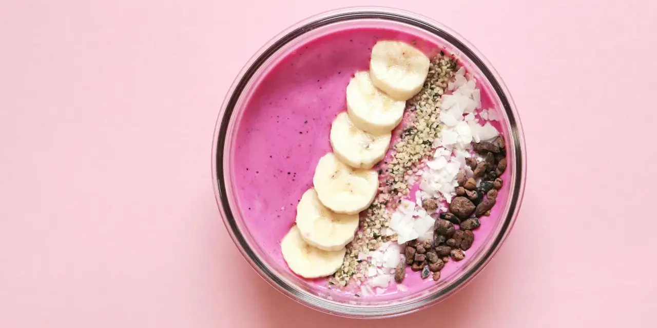 Un tazón de batido rosa vibrante con rodajas de plátano, coco rallado, semillas de chía y trozos de cacao.