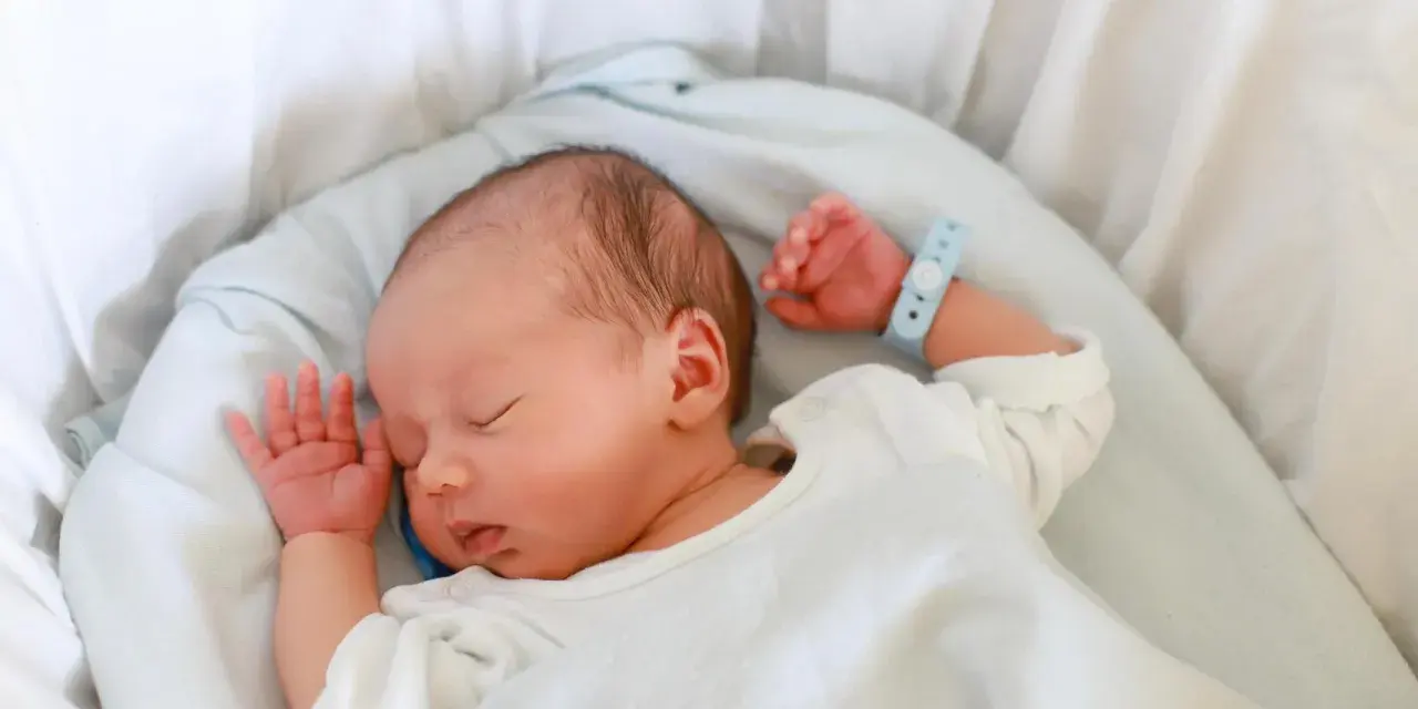Bebé recién nacido duerme plácidamente en su cuna. Su pulsera de identificación, con el **lua nombre**, indica que está seguro y cuidado.
