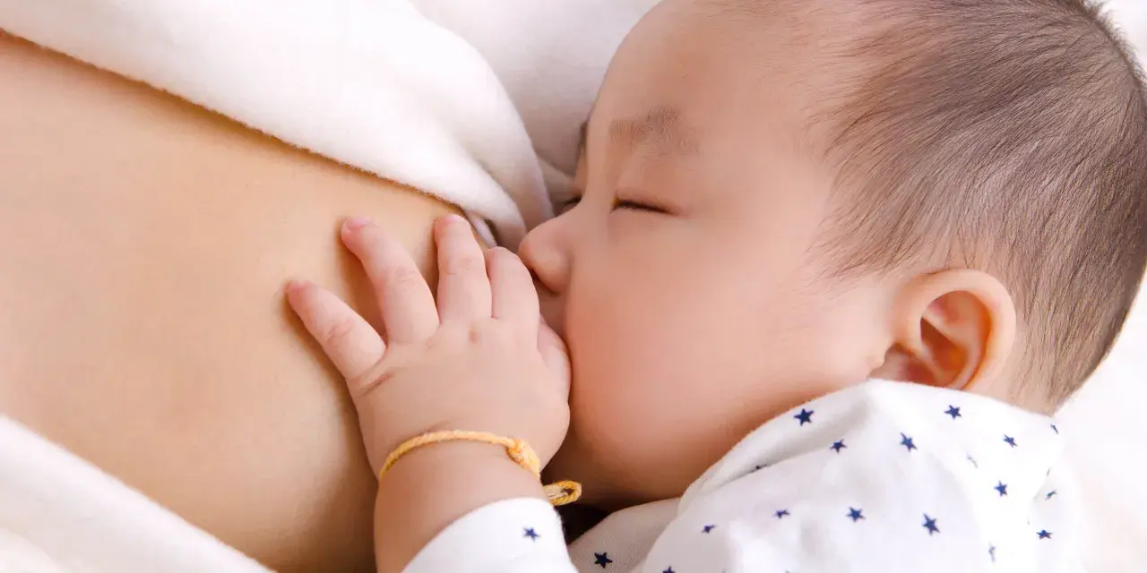 Bebé asiático durmiendo mientras se alimenta, mostrando el reflejo de succión. Su mano pequeña descansa sobre el pecho de su madre.