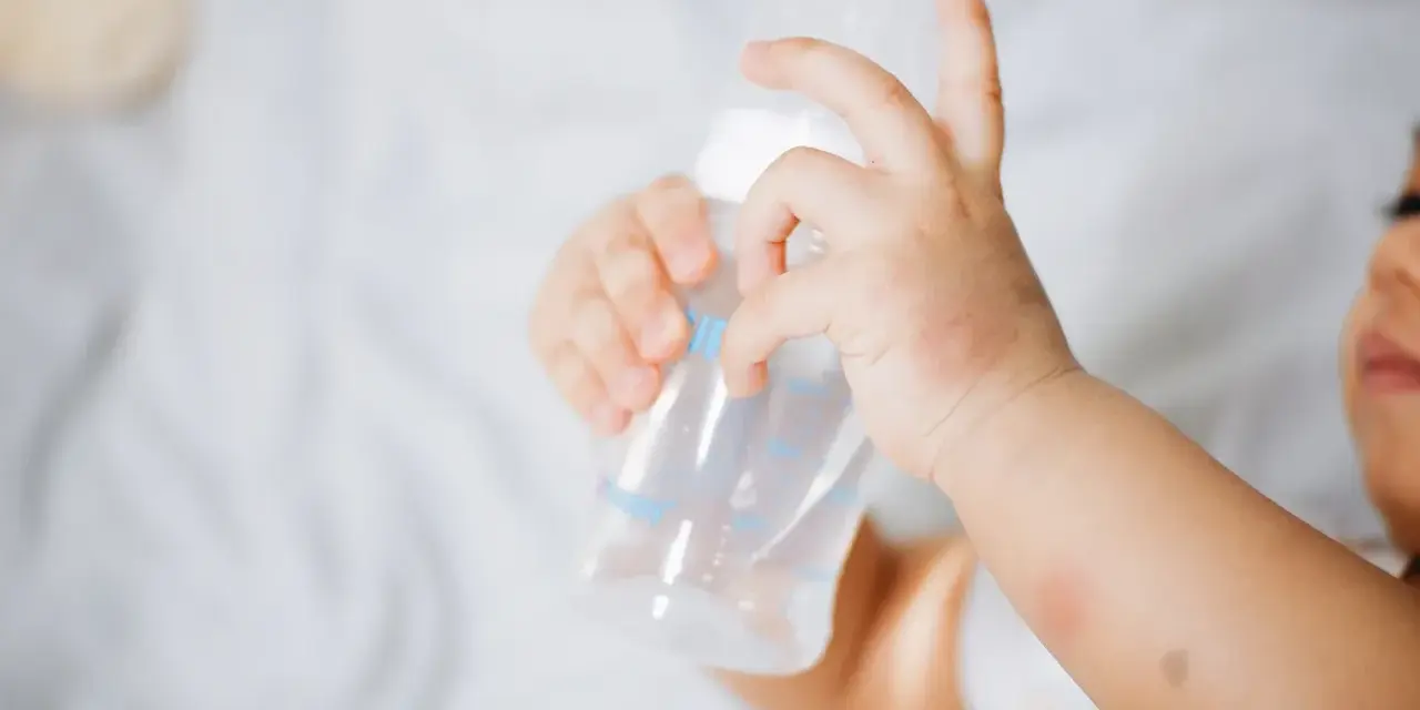 Manitas de bebé sujetando un biberón. Se aprecian rojeces en su piel, quizás por dermatitis o por el calor del sudor.