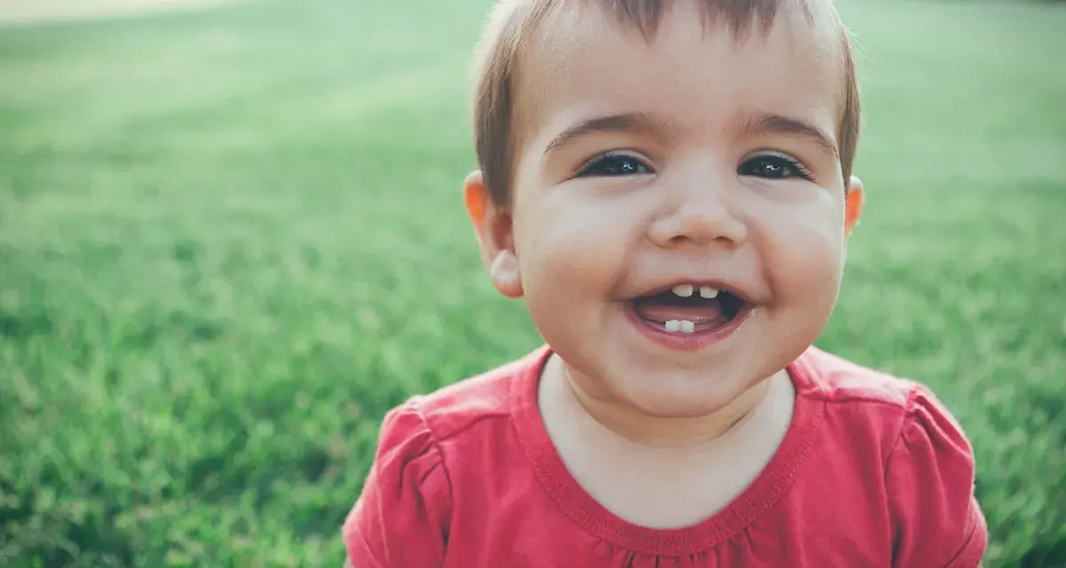 Un bebé de 1 año sin dientes ríe a carcajadas en el césped, mostrando dos pequeños dientes inferiores.