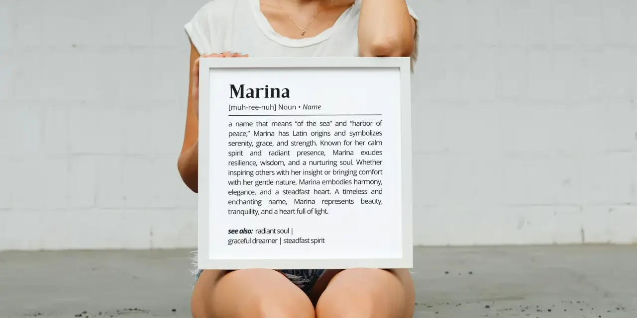 Una mujer sostiene un marco blanco con la definición del nombre Marina, que significa "del mar" y "puerto de paz".