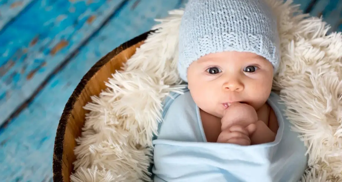 Bebé Ezra, con gorrito azul y envuelto en mantita, mira curioso. Descansa en una cesta sobre fondo de madera azul.