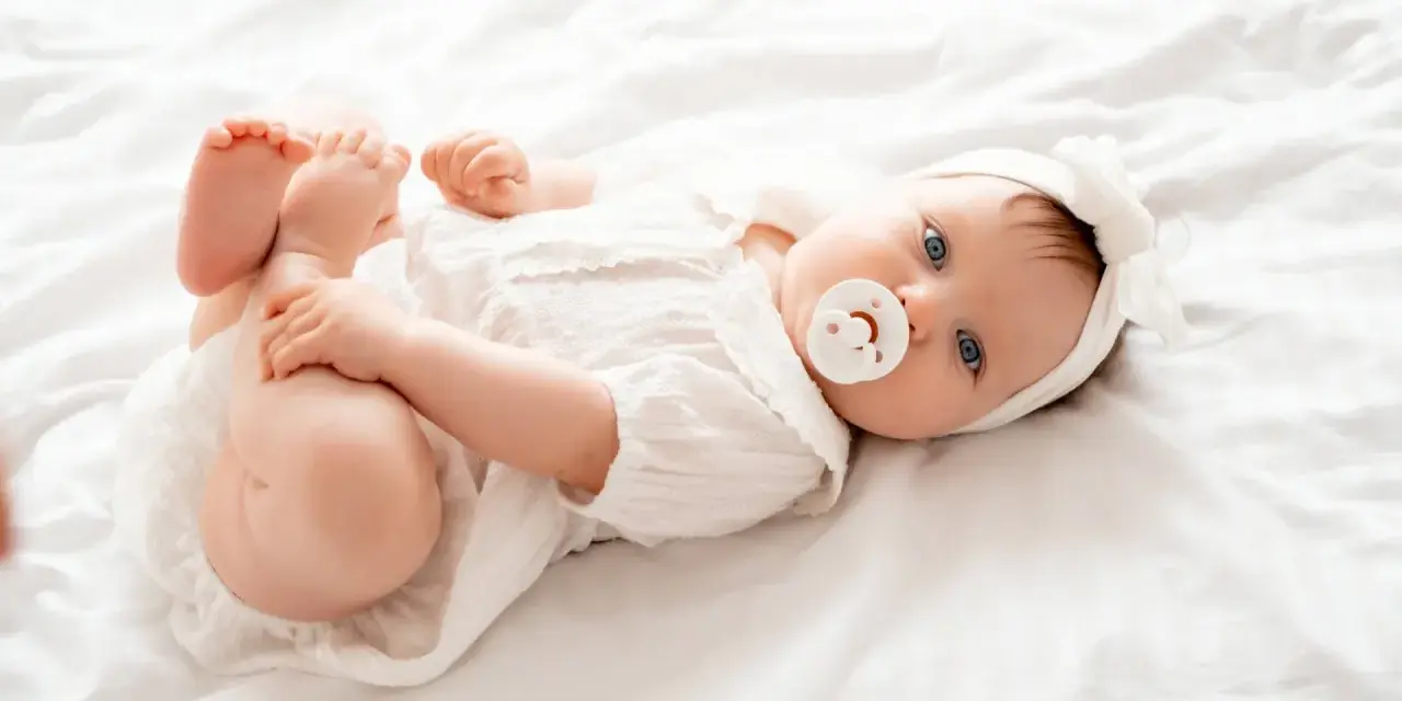 Bebé Ariadna, con su chupete y diadema blanca, juega con sus pies sobre una cama blanca.