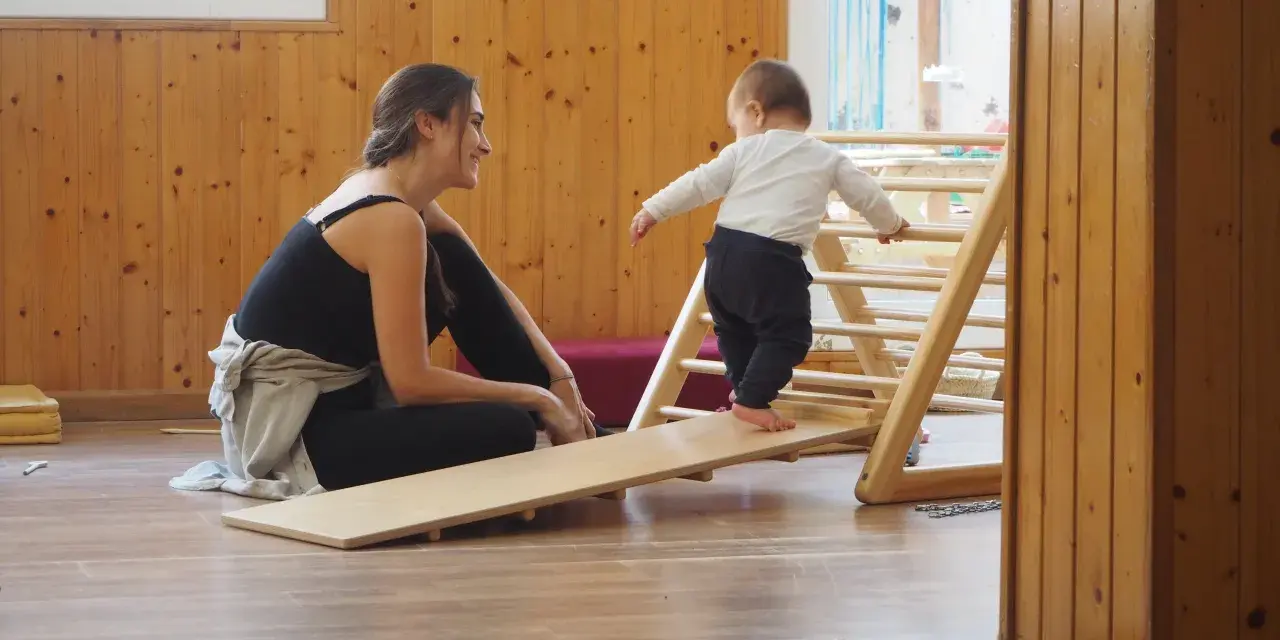 Niña gateando en rampa de madera, animada por mujer sonriente. Fomentando el movimiento libre y la exploración.