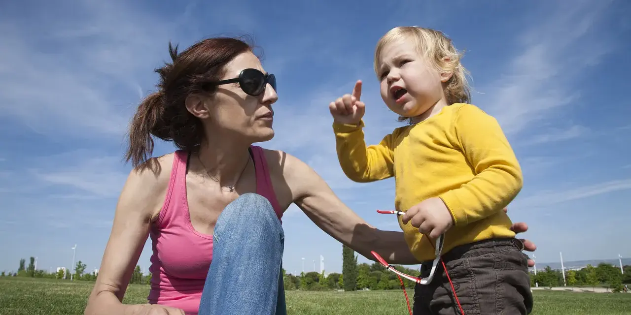 Niña rubia con camiseta amarilla, con el dedo levantado, parece que está aprendiendo cuando empiezan a balbucear los bebes. Madre con gafas de sol la mira.