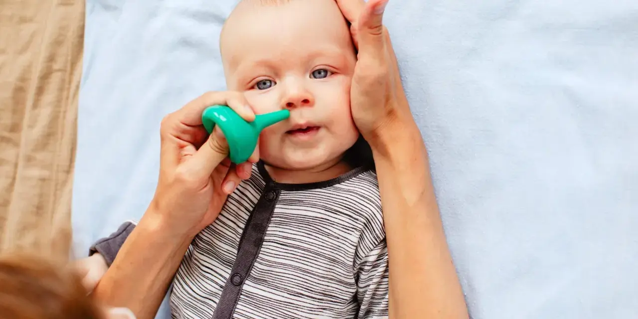 Bebé con ojos azules siendo ayudado a quitar mocos con un aspirador nasal verde.