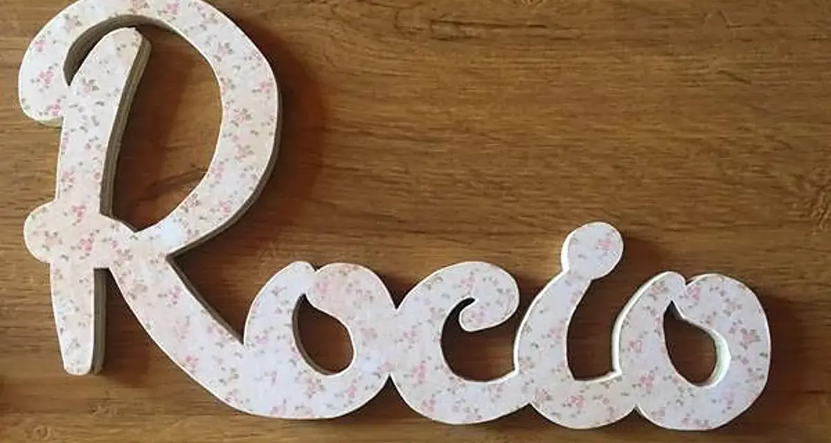 Letra decorativa con el nombre Rocio, cubierta de un delicado estampado floral rosa sobre fondo blanco, sobre una superficie de madera.