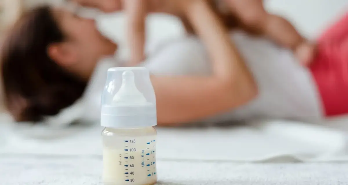 Biberón con leche, mientras una madre juega con su bebé. La imagen sugiere la pregunta: ¿cuanta leche toma un bebé de 1 mes?