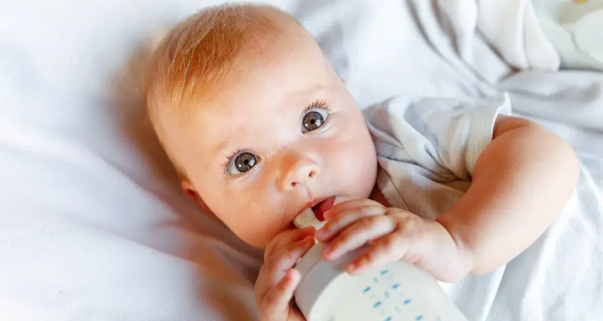 Bebé con ojos grandes y curiosos, bebiendo leche de un biberón. La imagen responde a la pregunta: cuanta leche toma un bebe.