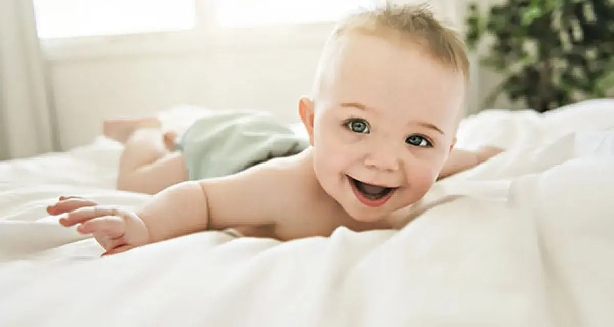 Bebé sonriente boca abajo en una cama blanca, con un reflejo de hociqueo en su expresión juguetona.