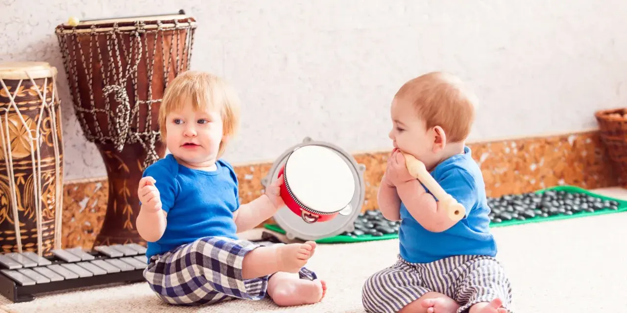 Dos bebés disfrutan de la estimulación musical temprana, uno con un tambor y otro con una flauta, rodeados de instrumentos.