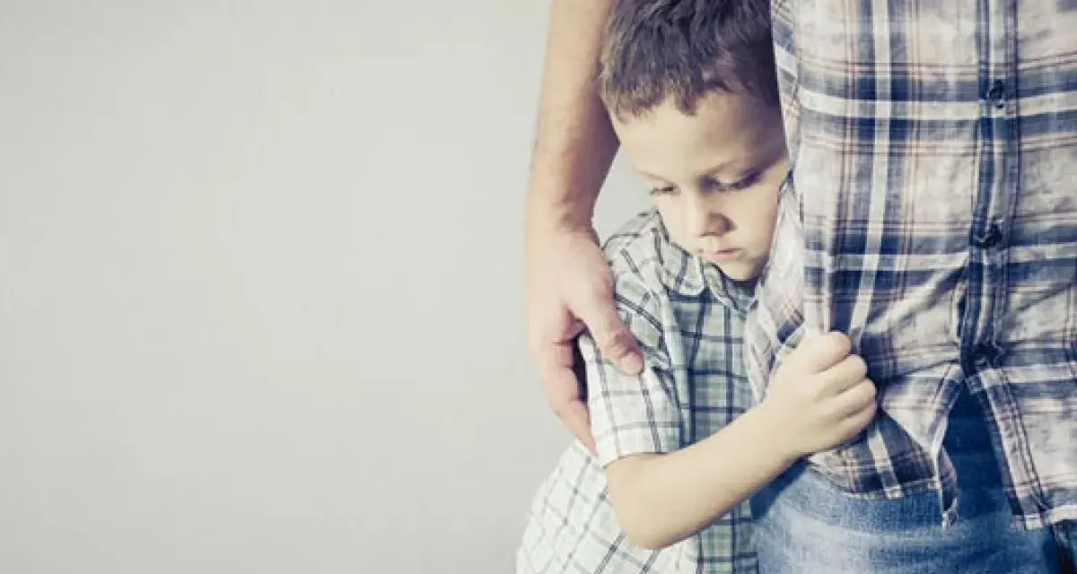 Un niño se aferra a la pierna de un adulto, con la cabeza baja, reflejando una **crisis** de **8 meses**.