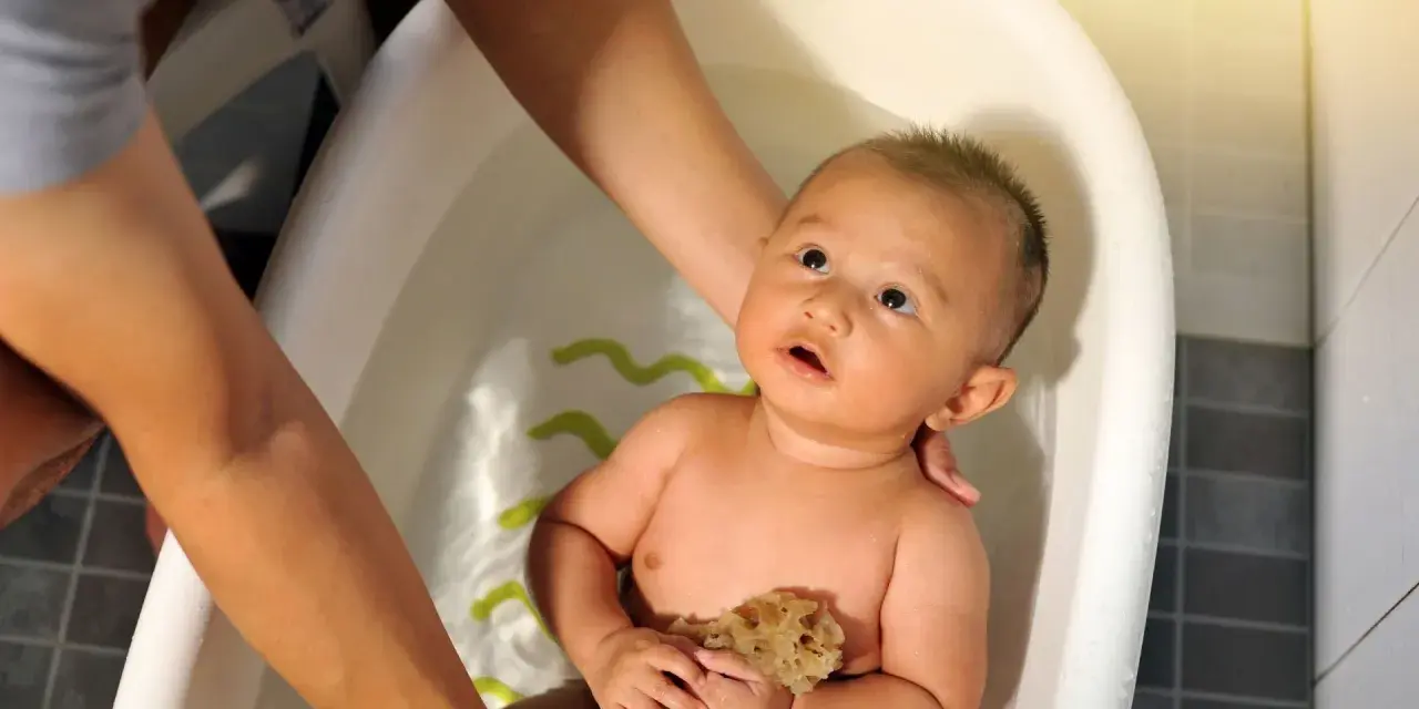 Bebé en bañera, manos adultas ajustan la temperatura del agua. Sostiene una esponja, mirando hacia arriba con curiosidad.