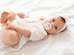 Bebé Ariadna, con su chupete y diadema blanca, juega con sus pies sobre una cama blanca.