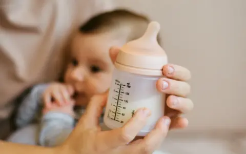 Manos sostienen un biberón con leche, con un bebé borroso al fondo. Es hora de considerar cuando cambiar tetina biberon.