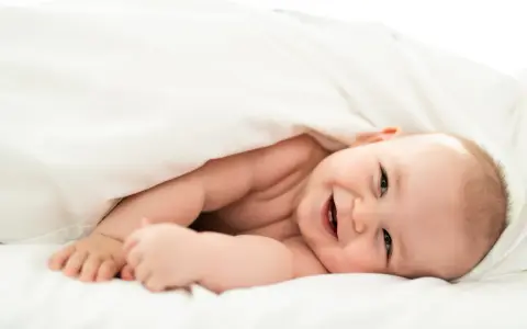 Un bebé sonriente se asoma bajo una manta blanca. Parece que los bebes tienen memoria, ¡y este ya recuerda cómo sonreír!