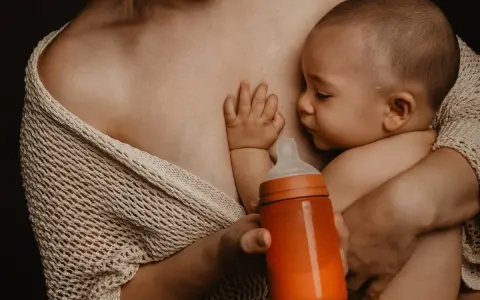 Bebé succionando un biberón naranja, con la mano apoyada en el pecho de la madre. Se aprecian varios tipos de tetinas en el mercado.