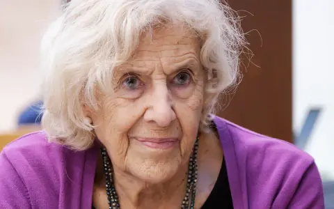 Retrato de una anciana con cabello blanco y ojos azules, luciendo un suéter morado. Su rostro arrugado evoca la sabiduría y el significado de una vida plena, como el nombre Manuela.