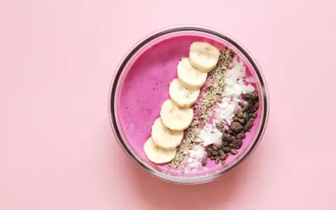 Un tazón de batido rosa vibrante con rodajas de plátano, coco rallado, semillas de chía y trozos de cacao.