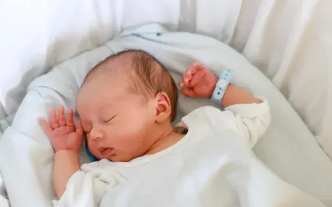 Bebé recién nacido duerme plácidamente en su cuna. Su pulsera de identificación, con el **lua nombre**, indica que está seguro y cuidado.