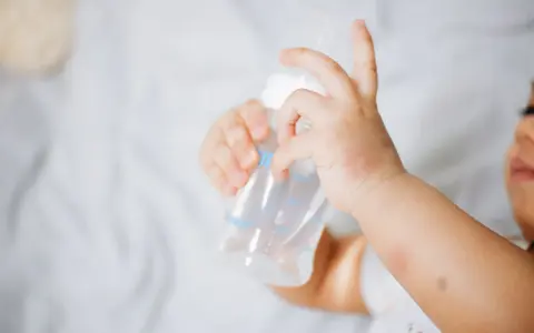 Manitas de bebé sujetando un biberón. Se aprecian rojeces en su piel, quizás por dermatitis o por el calor del sudor.