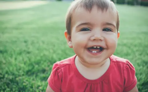 Un bebé de 1 año sin dientes ríe a carcajadas en el césped, mostrando dos pequeños dientes inferiores.
