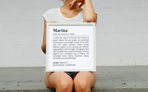 Una mujer sostiene un marco blanco con la definición del nombre Marina, que significa "del mar" y "puerto de paz".