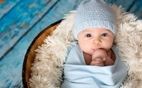 Bebé Ezra, con gorrito azul y envuelto en mantita, mira curioso. Descansa en una cesta sobre fondo de madera azul.