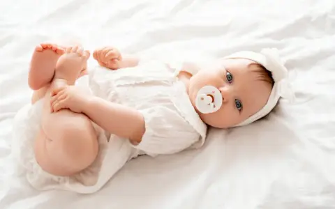 Bebé Ariadna, con su chupete y diadema blanca, juega con sus pies sobre una cama blanca.