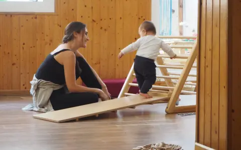 Niña gateando en rampa de madera, animada por mujer sonriente. Fomentando el movimiento libre y la exploración.
